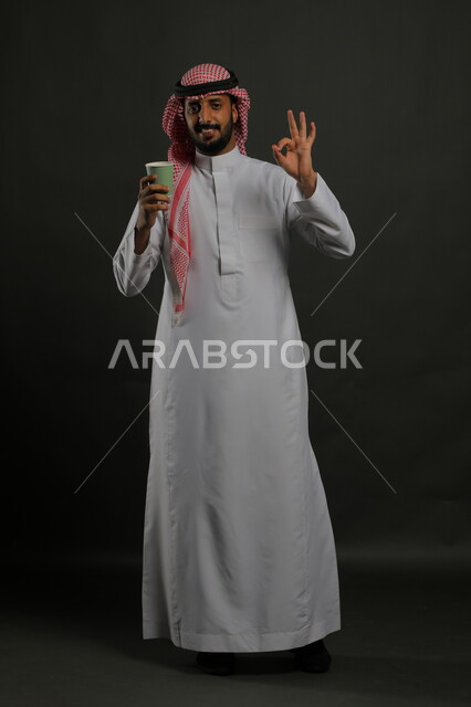 النشاط والحيوية عند شرب القهوة، النظر إلى الكاميرا بإيماءات الإعجاب والاستمتاع بتناول المشروبات الساخنة، بورتريه لرجل عربي سعودي مبتسم يرتدي الثوب التقليدي والشماغ يحمل بيده كوب ورقي ويشير  بعلامة الجودة والامتياز، صورة الجسم بالكامل، خليفية سوداء
