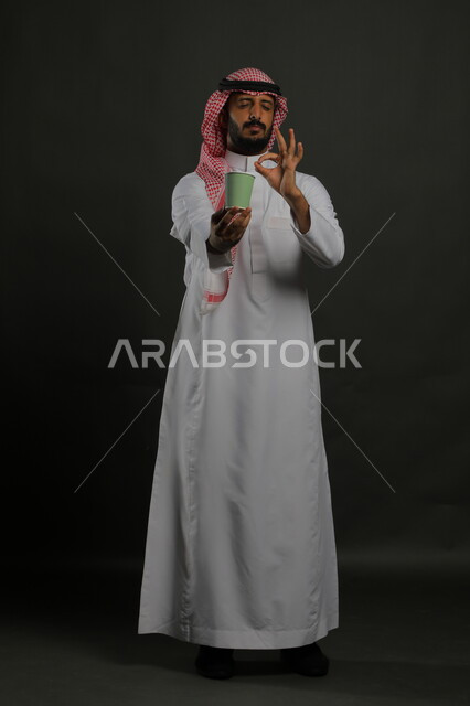 الحيوية والنشاط عند شرب القهوة، إيماءات الإعجاب والاستمتاع بتناول المشروبات الساخنة، بورتريه لرجل عربي سعودي يرتدي الثوب التقليدي والشماغ يحمل بيده كوب ورقي ويشير بعلامة الجودة والامتياز، صورة الجسم بالكامل، خليفية سوداء