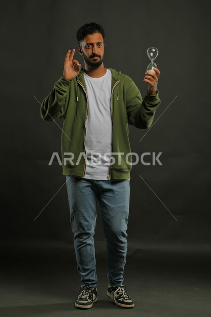 السرور بالقدرة على تنظيم الوقت، تحديد أوقات و مواعيد العمل، مفهوم إدارة الأعمال ،, بورتريه لرجل سعودي خليجي يرتدي الزي الكاجوال يحمل بيده الساعة الرملية الزجاجية (البنكام) يرفع يده بعلامو الجودة و ينظر إلى الكاميرا  ، صورة الجسم بالكامل، خلفية سوداء
