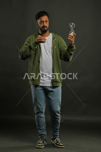 الإشارة إلى انقضاء الزمن بإيماءات الجدية والحزم، مفهوم إدارة الأعمال وتنظيم الأوقات، أدوات قديمه لتحديد الوقت, بورتريه لرجل عربي سعودي خليجي يرتدي الزي الكاجوال يحمل بيده الساعة الرملية الزجاجية (البنكام)، صورة الجسم بالطول الكامل، خلفية سوداء