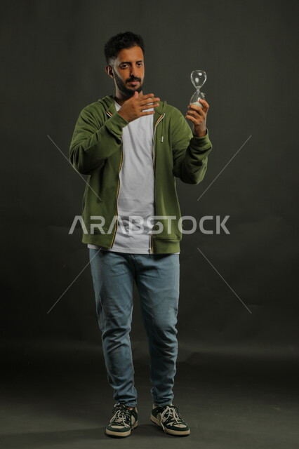 مفهوم تنظيم الأوقات و إدارة الأعمال، أدوات قديمه لتحديد الوقت, الإشارة إلى انقضاء الزمن بإيماءات الجدية والحزم، بورتريه لرجل عربي سعودي خليجي يرتدي الزي الكاجوال يحمل بيده الساعة الرملية الزجاجية (البنكام)، صورة الجسم بالطول الكامل، خلفية سوداء