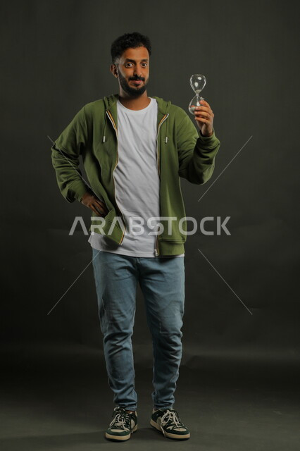 النظر إلى الكاميرا بإيماءات السرور، مفهوم إدارة الأعمال وتنظيم الجدول الزمني، أدوات قديمة لتحديد الوقت، القيام بمراقبة توقيت الرمل, بورتريه لرجل سعودي خليجي يرتدي الزي الكاجوال يحمل بيده الساعة الرملية الزجاجية (البنكام) ، صورة الجسم بالكامل، خلفية سوداء