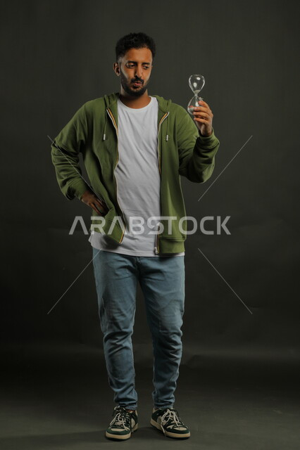 ايماءات وجه وفم تدل على الملل، مفهوم إدارة الأعمال وتنظيم الجدول الزمني، القيام بمراقبة توقيت الرمل, بورتريه لرجل سعودي خليجي يرتدي الزي الكاجوال يحمل بيده الساعة الرملية الزجاجية (البنكام) وينظر إليها بإيماءات الانتظار ، صورة الجسم بالكامل، خلفية سوداء