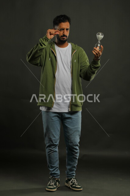 اشارات تدل على بقاء بعض الوقت، تحديد أوقات و مواعيد العمل، مفهوم إدارة الأعمال وتنظيم الجدول الزمني، القيام بمراقبة توقيت الرمل, بورتريه لرجل سعودي خليجي يرتدي الزي الكاجوال يحمل بيده الساعة الرملية الزجاجية (البنكام)، صورة الجسم بالكامل، خلفية سوداء