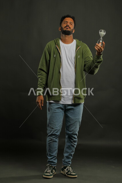 اغماض العينين و انتظار انقضاء الوقت، تحديد أوقات و مواعيد العمل، مفهوم إدارة الأعمال وتنظيم الجدول الزمني، القيام بمراقبة توقيت الرمل, بورتريه لرجل سعودي خليجي يرتدي الزي الكاجوال يحمل بيده الساعة الرملية الزجاجية (البنكام)، صورة الجسم بالكامل، خلفية سوداء