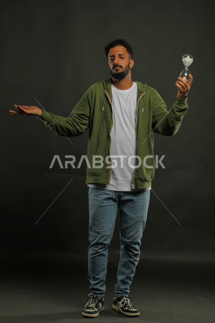ادارك قيمه اللحظات الثمينة في حياتنا ،أدوات قديمه لتحديد الوقت, بورتريه لرجل عربي سعودي خليجي يرتدي الزي الكاجوال يحمل الساعة الرملية الزجاجية (البنكام) يشير بيده بإيماءات الملل وعدم الصبر، إدارة الجدول الزمني, صورة الجسم بالطول الكامل، خلفية سوداء