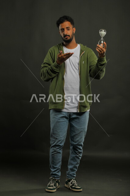 الإشارة إلى انقضاء الزمن بإيماءات الجدية والحزم، أدوات  قديمه لتحديد الوقت, بورتريه لرجل عربي سعودي خليجي يرتدي الزي الكاجوال يحمل بيده الساعة الرملية الزجاجية (البنكام)، إدارة الجدول الزمني, صورة الجسم بالطول الكامل، خلفية سوداء