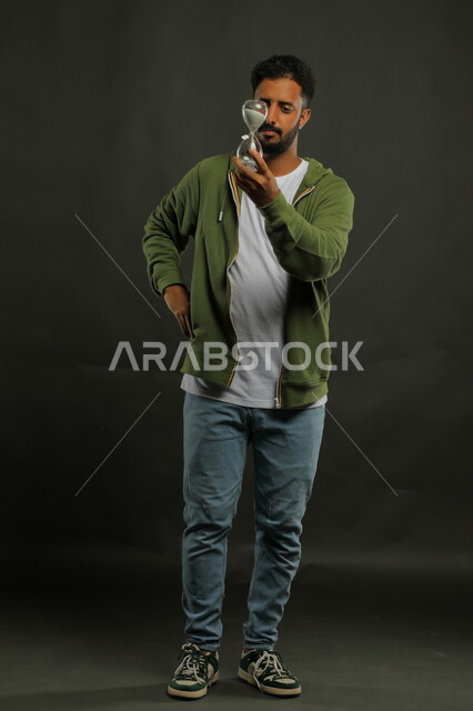 ادارك قيمة الوقت و اللحظات الثمينة في حياتنا ،الوقوف بتعابير الملل والانتظار، القيام بمراقبة توقيت الرمل, بورتريه لرجل سعودي خليجي يرتدي الزي الكاجوال يحمل بيده الساعة الرملية الزجاجية (البنكام) وينظر إليها بإيماءات الصبر، صورة الجسم بالكامل، خلفية سوداء