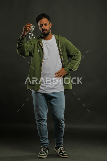 ادارك قيمة اللحظات الثمينة في حياتنا ،الوقوف بتعابير الملل والانتظار، القيام بمراقبة توقيت الرمل, بورتريه لرجل سعودي خليجي يرتدي الزي الكاجوال يحمل بيده الساعة الرملية الزجاجية (البنكام) وينظر إليها بإيماءات الصبر، صورة الجسم بالكامل، خلفية سوداء