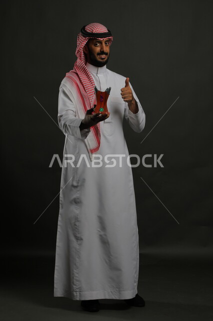 إيماءات الإعجاب برائحة العود والبخور الزكية, بورتريه لشاب عربي خليجي سعودي مبتسم يرتدي الشماغ والثوب التقليدي يحمل المبخرة ويرفع إصبعه الإبهام نحو الأعلى بعلامة الجودة, الأناقة والاهتمام بالمظهر الخارجي, صورة الجسم بالطول الكامل, خلفية رمادية