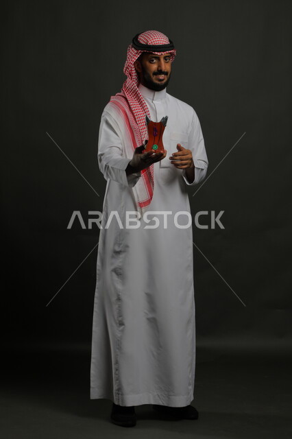 ايماءات الفرح بالأعياد والمناسبات, التطيب بالعود والبخور, بورتريه لشاب عربي خليجي سعودي مبتسم يرتدي الشماغ والثوب التقليدي ينظر إلى الكاميرا ويحمل المبخرة, الأناقة والاهتمام بالمظهر الخارجي, العادات والتقاليد السعودية, صورة الجسم بالطول الكامل, خلفية رمادية