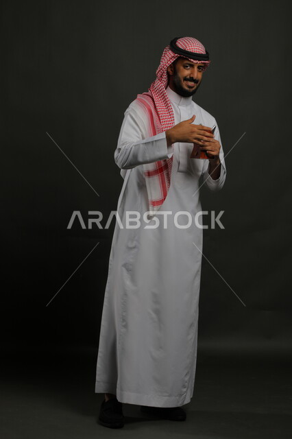 الأناقة والاهتمام بالمظهر الخارجي, التطيب بالعود والبخور, إيماءات الفرح بالأعياد والمناسبات, بورتريه لشاب عربي خليجي سعودي مبتسم يرتدي الشماغ والثوب التقليدي ينظر إلى الكاميرا ويحمل المبخرة,  صورة الجسم بالطول الكامل, خلفية رمادية