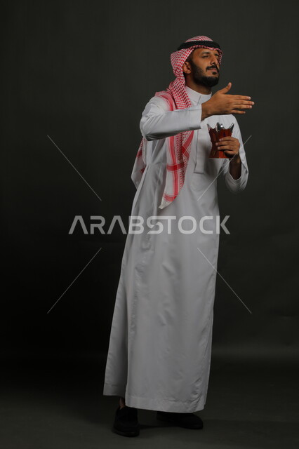 استنشاق رائحة العطور والبخور, العادات والتقاليد السعودية, استخدام خشب العود بالأعياد والمناسبات, بورتريه لشاب عربي خليجي سعودي يرتدي الشماغ والثوب التقليدي ويحمل المبخرة, الأناقة والاهتمام بالمظهر الخارجي, صورة الجسم بالطول الكامل, خلفية رمادية