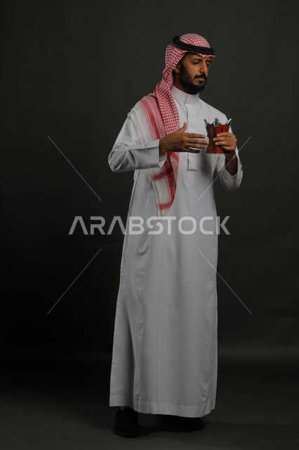  الاستمتاع برائحة العطور والبخور, العادات والتقاليد السعودية, استخدام خشب العود بالأعياد والمناسبات, بورتريه لشاب عربي خليجي سعودي يرتدي الشماغ والثوب التقليدي ويحمل المبخرة, الأناقة والاهتمام بالمظهر الخارجي, صورة الجسم بالطول الكامل, خلفية رمادية