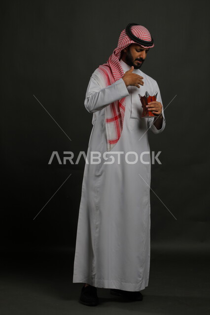 العادات والتقاليد السعودية, استخدام خشب العود بالأعياد والمناسبات, التطيب بالعطور والبخور, بورتريه لشاب عربي خليجي سعودي يرتدي الشماغ والثوب التقليدي ويحمل المبخرة, الأناقة والاهتمام بالمظهر الخارجي, صورة الجسم بالطول الكامل, خلفية رمادية