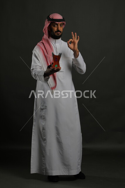 الإعجاب برائحة العود الزكية, ايماءات علامة جودة البخور السعودي، منتج محلي وطني, بورتريه لشاب عربي خليجي سعودي مبتسم يرتدي الشماغ والثوب التقليدي يحمل المبخرة, الأناقة والاهتمام بالمظهر الخارجي, صورة الجسم بالطول الكامل, خلفية رمادية