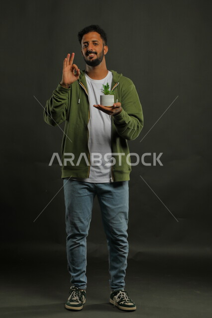 إيماءات علامة الجوده والإعجاب وحب النباتات والاهتمام بها, بورتريه لشاب عربي خليجي سعودي مبتسم يرتدي الزي الكاجوال يقف باستقامة ويحمل بيده حوض زراعي صغير، العناية بالشتلات المنزلية، صورة الجسم بالطول الكامل، خلفية سوداء