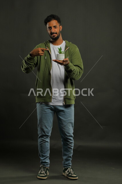 تقديم النصيحة لاختيار نوع الزهرة, النظر إلى الكاميرا والإشارة إلى النبتة الصغيرة, بورتريه لشاب عربي خليجي سعودي يرتدي الزي الكاجوال يحمل بيده حوض زراعي، حب النباتات والاهتمام بها، العناية بالشتلات المنزلية، صورة الجسم بالطول الكامل، خلفية سوداء