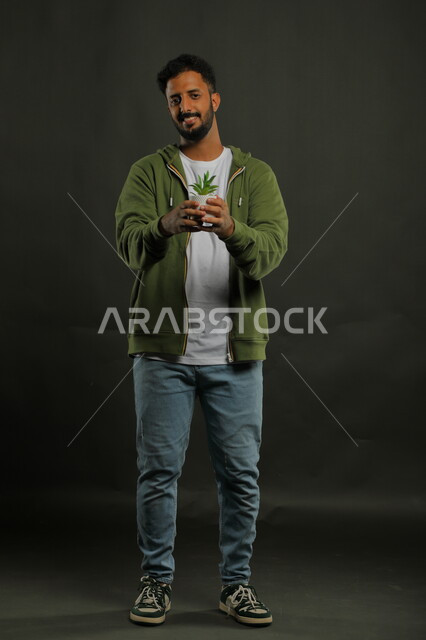 العناية بنباتات الزينة, الاهتمام بالشتلات المنزلية, بورتريه لرجل عربي خليجي سعودي مبتسم يرتدي الزي الكاجوال يحمل بيده حوض زراعي صغير, الوقوف باستقامة والنظر إلى الكاميرا بإيماءات السرور, صورة الجسم بالطول الكامل, خلفية سوداء