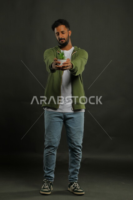 العناية بنباتات الزينة, الاهتمام بالشتلات المنزلية, بورتريه لرجل عربي خليجي سعودي يرتدي الزي الكاجوال يحمل بيده حوض زراعي صغير, الوقوف باستقامة والنظر إلى الكاميرا بإيماءات السرور, صورة الجسم بالطول الكامل, خلفية سوداء
