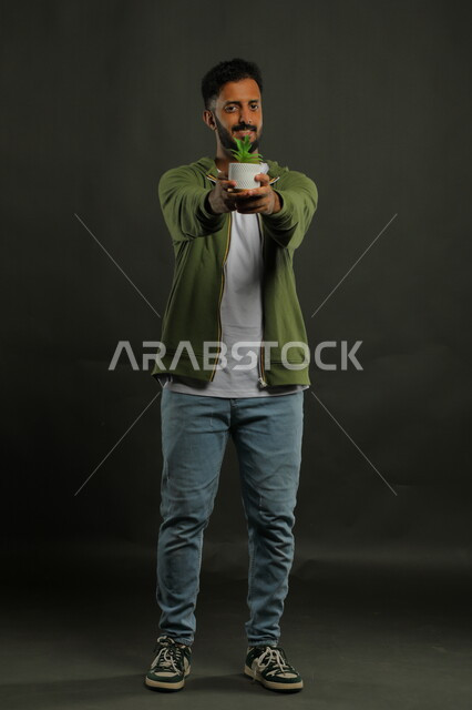 العناية بنباتات الزينة, الاهتمام بالشتلات المنزلية, بورتريه لرجل عربي خليجي سعودي مبتسم يرتدي الزي الكاجوال يحمل بيده حوض زراعي صغير, الوقوف باستقامة والنظر إلى الكاميرا بإيماءات السرور, صورة الجسم بالطول الكامل, خلفية سوداء