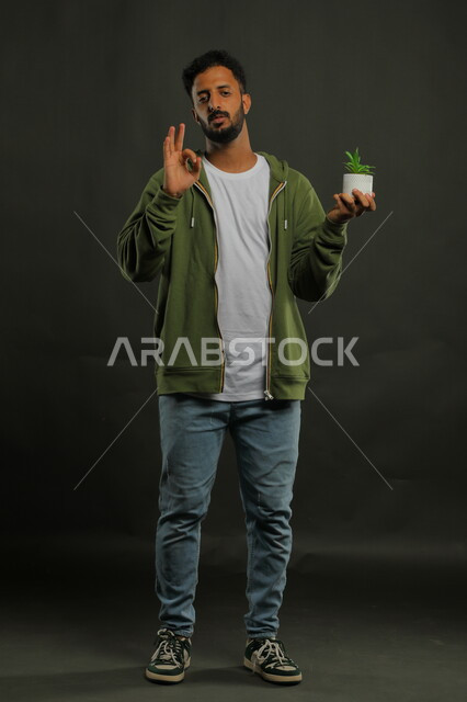 إيماءات علامة الجوده والإعجاب وحب النباتات والاهتمام بها, بورتريه لشاب عربي خليجي سعودي يرتدي الزي الكاجوال يقف باستقامة ويحمل بيده حوض زراعي صغير، العناية بالشتلات المنزلية، صورة الجسم بالطول الكامل، خلفية سوداء
