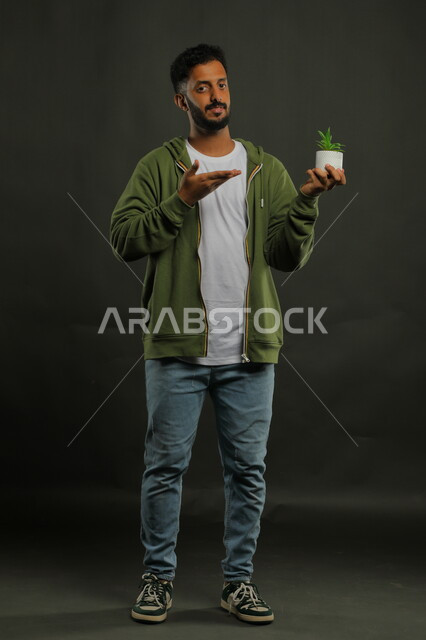 تقديم النصيحة لاختيار نوع الزهرة, النظر إلى الكاميرا والإشارة إلى النبتة الصغيرة, بورتريه لشاب عربي خليجي سعودي يرتدي الزي الكاجوال يحمل بيده حوض زراعي، حب النباتات والاهتمام بها، العناية بالشتلات المنزلية، صورة الجسم بالطول الكامل، خلفية سوداء