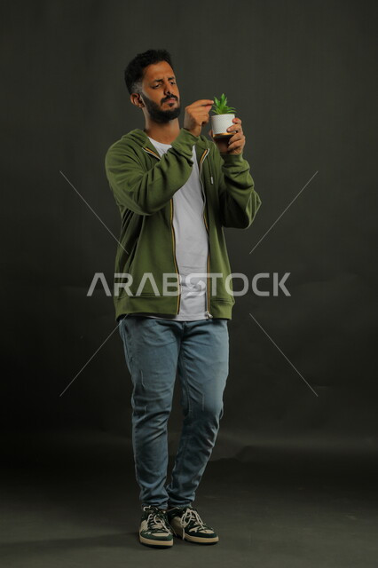 تحديد واكتشاف نوع الزهرة, الوقوف باستقامة بإيماءات التركيز والدقة, بورتريه لشاب عربي خليجي سعودي يرتدي الزي الكاجوال ويحمل بيده حوض زراعي صغير، حب النباتات والاهتمام بها، العناية بالشتلات المنزلية، صورة الجسم بالطول الكامل، خلفية سوداء