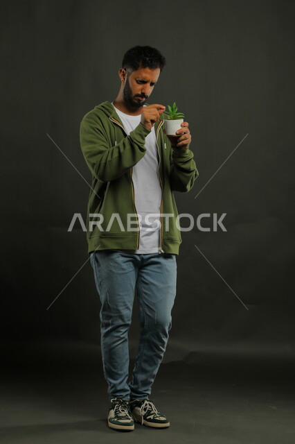 تحديد واكتشاف نوع الزهرة, الوقوف باستقامة بإيماءات التركيز والدقة, بورتريه لشاب عربي خليجي سعودي يرتدي الزي الكاجوال ويحمل بيده حوض زراعي صغير، حب النباتات والاهتمام بها، العناية بالشتلات المنزلية، صورة الجسم بالطول الكامل، خلفية سوداء