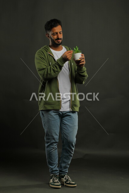 Discovering and identifying the type of flower, standing up straight with gestures of focus and precision, portrait of a Saudi Gulf Arab young man wearing casual clothes and holding a small agricultural basin in his hand, love and care for plants, caring for home seedlings, full-length body portrait, black background