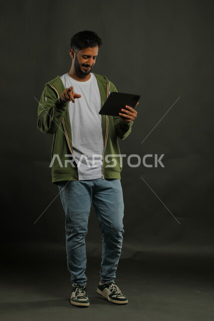إجراء مكالمة فيديو مع الاصدقاء بإيماءات السعادة والسرور, متابعة آخر أخبار السوشال ميديا, بورتريه لشاب عربي خليجي سعودي مبتسم  يرتدي الزي الكاجوال يستخدم الجهاز اللوحي يشير بيده إلى الأمام, صورة الجسم بالكامل, خلفية سوداء