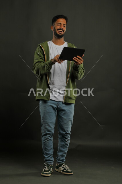 التسامر والضحك مع الأهل والأصدقاء أثناء إجراء مكالمة فيديو, متابعة آخر أخبار السوشال ميديا, بورتريه لشاب عربي خليجي سعودي مبتسم  يرتدي الزي الكاجوال يستخدم جهاز التابلت بإيماءات السرور, استخدام التكنولوجيا والتقنيات الحديثة، صورة الجسم بالكامل, خلفية سوداء