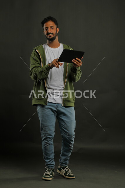 التسويق الالكتروني الذكي والآمن عبر الانترنت, استخدام تطبيقات وتقنيات التكنولوجيا والأجهزة الحديثة, بورتريه لشاب عربي خليجي سعودي مبتسم يرتدي الزي الكاجوال يحمل الجهاز اللوحي, الدردشة والتواصل مع الاصدقاء, صورة الجسم بالطول الكامل, خلفية سوداء