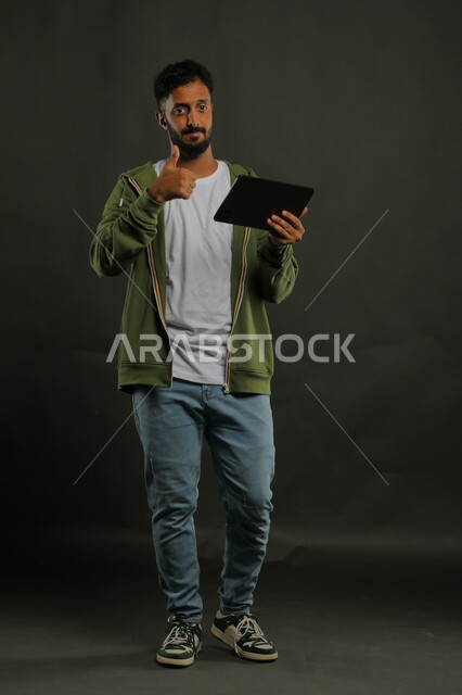 ايماءات القبول والرضا، دمج التكنولوجيا في الحياة العملية، بورتريه لشاب عربي خليجي سعودي مبتسم يرتدي الزي الكاجوال يرفع اصبع الابهام للأعلى بعلامة الجودة و ينظر إلى الكاميرا، تصفح مواقع التواصل الاجتماعي عبر جهاز التابليت، صورة الجسم بالكامل، خلفية سوداء