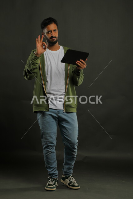  دمج التكنولوجيا في الحياة العملية، ايماءات الإعجاب والرضا، بورتريه لشاب عربي خليجي سعودي يرتدي الزي الكاجوال يشير بعلامة الجودة والامتياز و ينظر إلى الكاميرا، تصفح مواقع التواصل الاجتماعي عبر جهاز التابليت، صورة الجسم بالكامل، خلفية سوداء