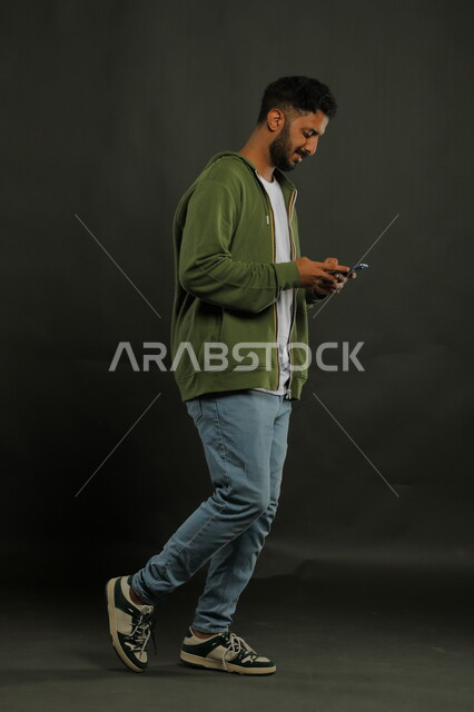 التواصل مع الأصدقاء وتبادل الأحاديث الودية عبر الجوال بإيماءات السرور, مفهوم صلة الرحم, بورتريه لشاب عربي خليجي سعودي مبتسم يرتدي الزي الكاجوال يقوم بتصفح مواقع السوشال ميديا عبر الهاتف المحمول, صورة الجسم بالطول الكامل, خلفية سوداء