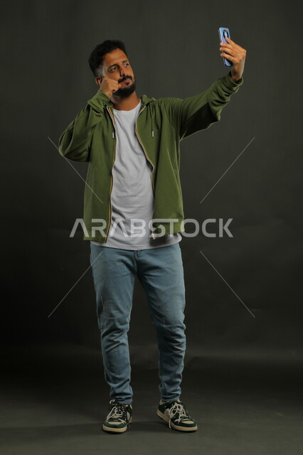 Making a video call via mobile phone, portrait of a Saudi Gulf Arab young man wearing casual clothes broadcasting live via social networking sites, using mobile phone applications and technologies, full-length body photo, black background