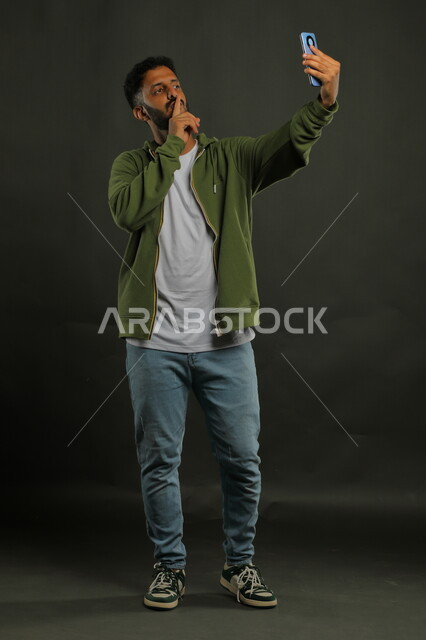 إجراء مكالمة فيديو عبر الجوال, بورتريه لشاب عربي خليجي سعودي يرتدي الزي الكاجوال يقوم بالبث المباشر عبر مواقع التواصل الاجتماعي, استخدام تطبيقات وتقنيات الهاتف المحمول, صورة الجسم بالطول الكامل, خلفية سوداء