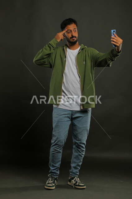 إجراء مكالمة فيديو عبر الجوال, بورتريه لشاب عربي خليجي سعودي يرتدي الزي الكاجوال يقوم بالبث المباشر عبر مواقع التواصل الاجتماعي, استخدام تطبيقات وتقنيات الهاتف المحمول, صورة الجسم بالطول الكامل, خلفية سوداء