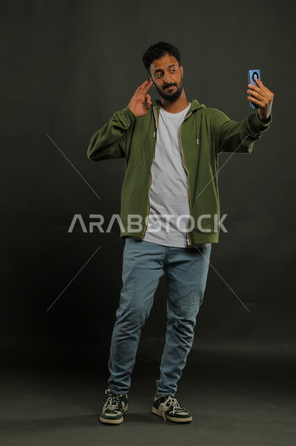 إجراء مكالمة فيديو عبر الجوال,  توثيق اللحظات السعيدة, بورتريه لشاب عربي خليجي سعودي يرتدي الزي الكاجوال يقوم التقاط صور سيلفي تذكارية عبر الهاتف المحمول, صورة الجسم بالطول الكامل, خلفية سوداء