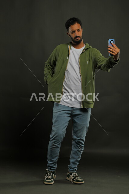 التقاط صور سيلفي تذكارية عبر الهاتف المحمول, توثيق اللحظات السعيدة, بورتريه لشاب عربي خليجي سعودي يرتدي الزي الكاجوال يقوم بإجراء مكالمة فيديو عبر الجوال, صورة الجسم بالطول الكامل, خلفية سوداء
