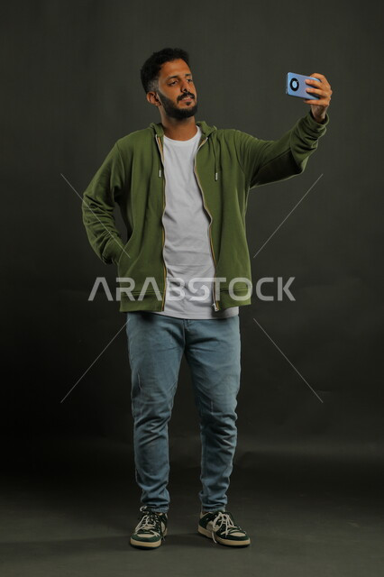 التقاط صور سيلفي تذكارية عبر الهاتف المحمول, توثيق اللحظات السعيدة, بورتريه لشاب عربي خليجي سعودي يرتدي الزي الكاجوال يقوم بإجراء مكالمة فيديو عبر الجوال, صورة الجسم بالطول الكامل, خلفية سوداء
