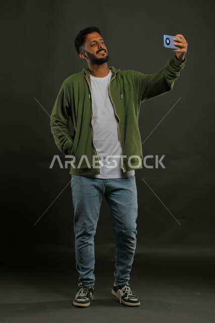 التقاط صور سيلفي تذكارية عبر الهاتف المحمول, توثيق اللحظات السعيدة, بورتريه لشاب عربي خليجي سعودي يرتدي الزي الكاجوال يقوم بإجراء مكالمة فيديو عبر الجوال, صورة الجسم بالطول الكامل, خلفية سوداء