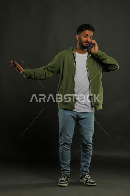 رفع اليد والشعور بالرفض وطلب عدم التدخل, ايماءات المنع والتوقف عن فعل شيء ما, بورتريه لشاب عربي خليجي سعودي يرتدي الزي الكاجوال يقوم بإجراء مكالمة صوتية عبر الهاتف المحمول بانزعاج وعبوس, صورة الجسم بالطول الكامل, خلفية سوداء