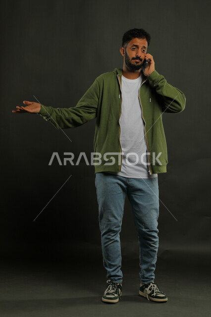 استخدام الأجهزة والتقنيات الحديثة, الاستفسار والسؤال عن أمر معين بإيماءات الاستفهام, بورتريه لشاب عربي خليجي سعودي يرتدي الزي الكاجوال يقوم بإجراء مكالمة هاتفية عبر الهاتف المحمول, صورة الجسم بالطول الكامل, خلفية سوداء