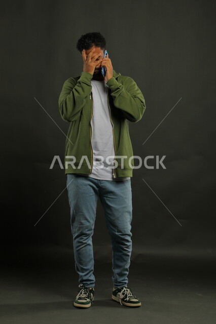 Stress, anxiety and feeling of disappointment, portrait of a Saudi Gulf Arab young man wearing casual clothes making a voice call via mobile phone with gestures of annoyance and bewilderment at hearing bad news, using modern and advanced communication devices, full-length body image, black background