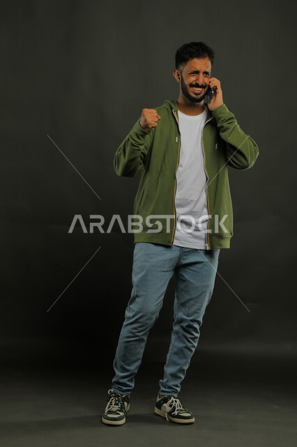 البشاشة واالسرور لربح الفريق المفضل, التواصل مع الأصدقاء وتبادل الأحاديث الودية عبر الجوال, بورتريه لشاب عربي خليجي سعودي مبتسم يرتدي الزي الكاجوال يقوم بإجراء مكالمة صوتية عبر الهاتف المحمول بإيماءات تحقيق الفوز والانتصار, صورة الجسم بالكامل, خلفية سوداء