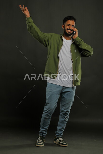تبادل الأحاديث الودية مع الأصدقاء عبر الجوال, بورتريه لشاب عربي خليجي سعودي مبتسم يرتدي الزي الكاجوال يقوم بإجراء مكالمة صوتية عبر الهاتف المحمول, مشاعر الاطمئنان والسؤال عن الحال, البشاشة والسرور لسماع الأخبار الجيده, صورة الجسم بالكامل, خلفية سوداء