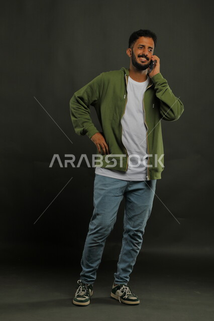 التواصل مع الأصدقاء وتبادل الأحاديث الودية عبر الجوال, مفهوم صلة الرحم, بورتريه لشاب عربي خليجي سعودي مبتسم يرتدي الزي الكاجوال يقوم بإجراء مكالمة صوتية عبر الهاتف المحمول, صورة الجسم بالكامل, خلفية سوداء