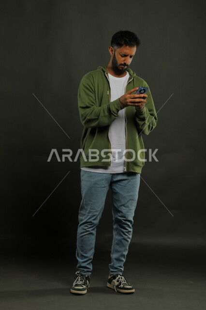 دردشات ومحادثات مع الأصدقاء عبر الأونلاين, بورتريه لشاب عربي خليجي سعودي يرتدي الزي الكاجوال يقوم باستخدام الهاتف المحمول بايماءات الانزعاج والعبوس, القلق والحزن لسماع الأخبار السيئة, صورة الجسم بالكامل, خلفية سوداء